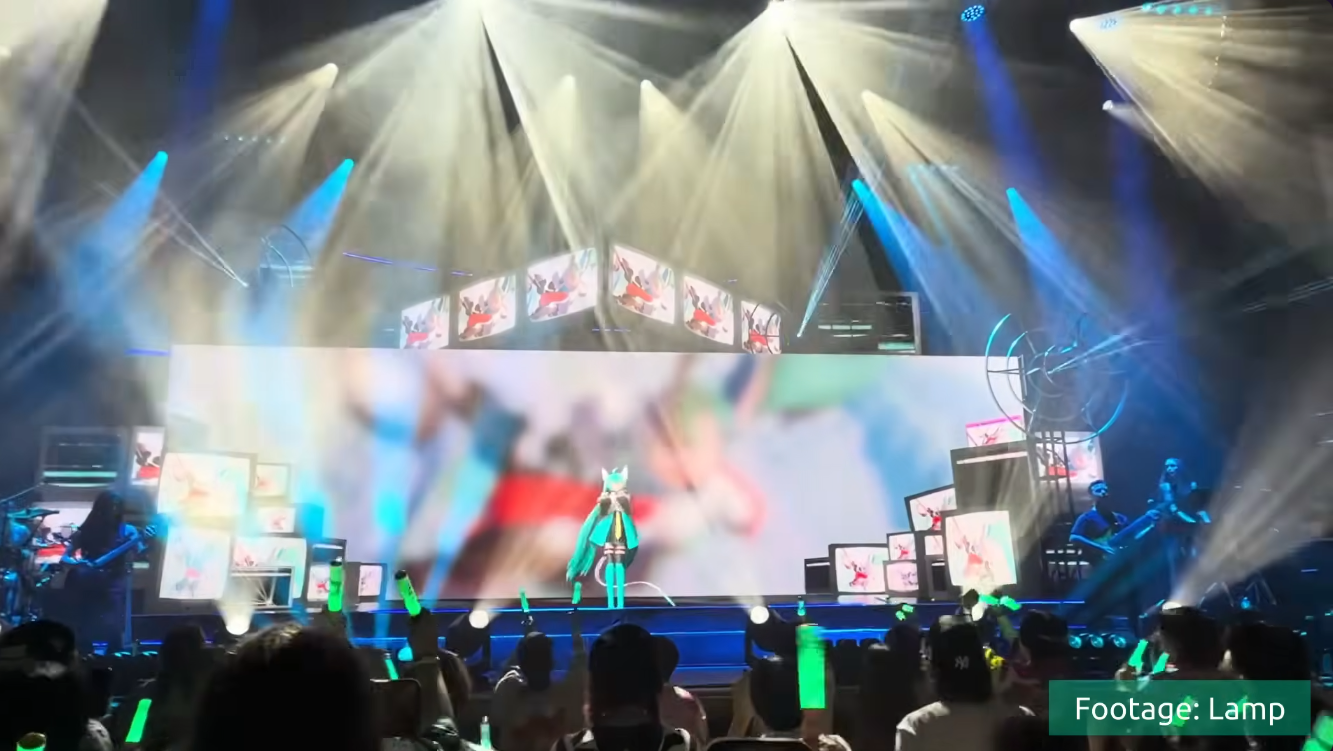 Miku on stage in front of a wall of old TVs on the wide LED screen, with the band beside her